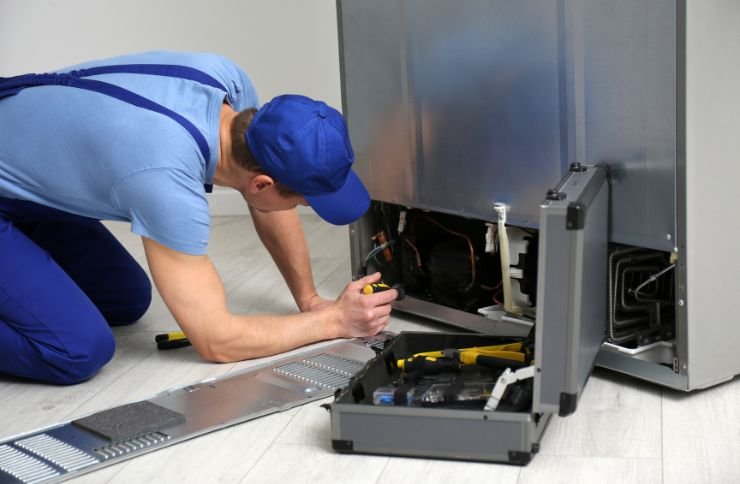 A man in blue overalls is seen repairing a refrigerator, illustrating commercial appliance repair services in Las Vegas, NV.