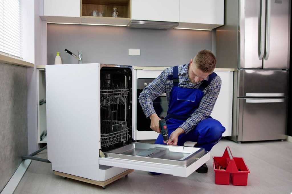 A man in blue overalls repairs a dishwasher, focused on the task at hand in a well-lit kitchen environment.